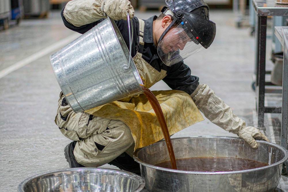 worker in protective gear pouring viscous amber epoxy resin into a steel mixing container in a workshop