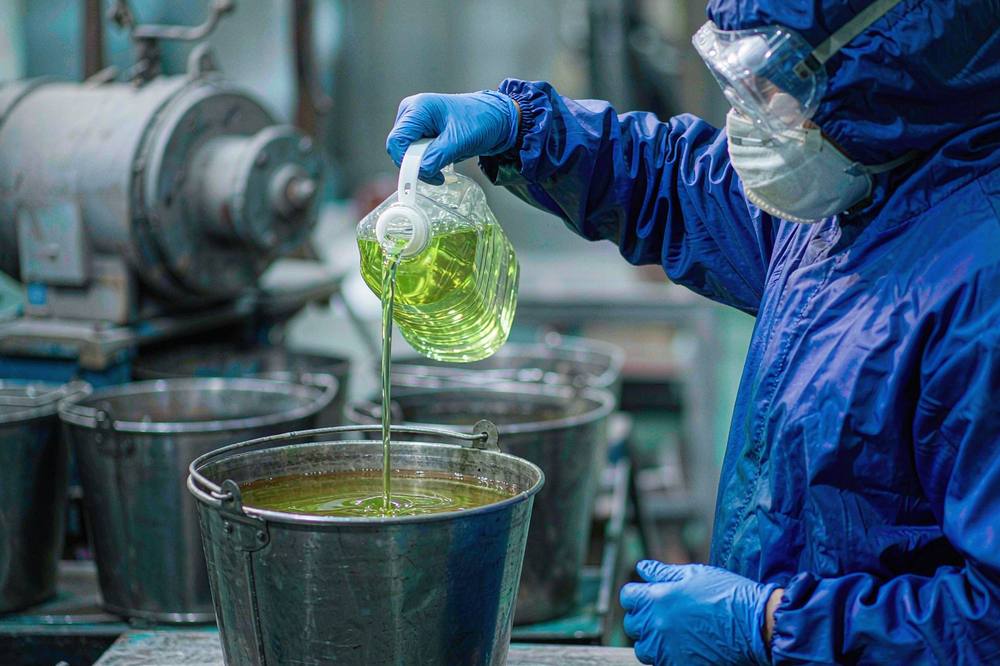 worker pouring pale amber cardanol diglycidyl ether diluent from a steel jug into an epoxy resin mixing pail