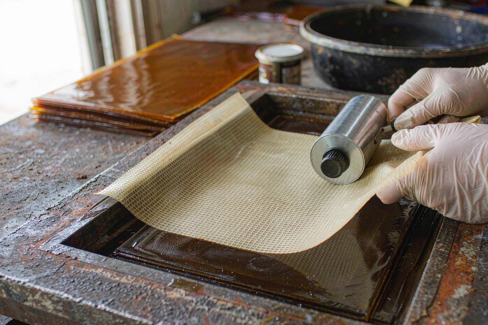gloved hands wetting out woven glass fibre fabric with amber novolac epoxy resin using a roller, cured laminate sheets stacked behind